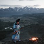 Full length of serious Tibetan male in traditional wear looking at campfire while standing with lute against mountains with cloudy sky at sunset