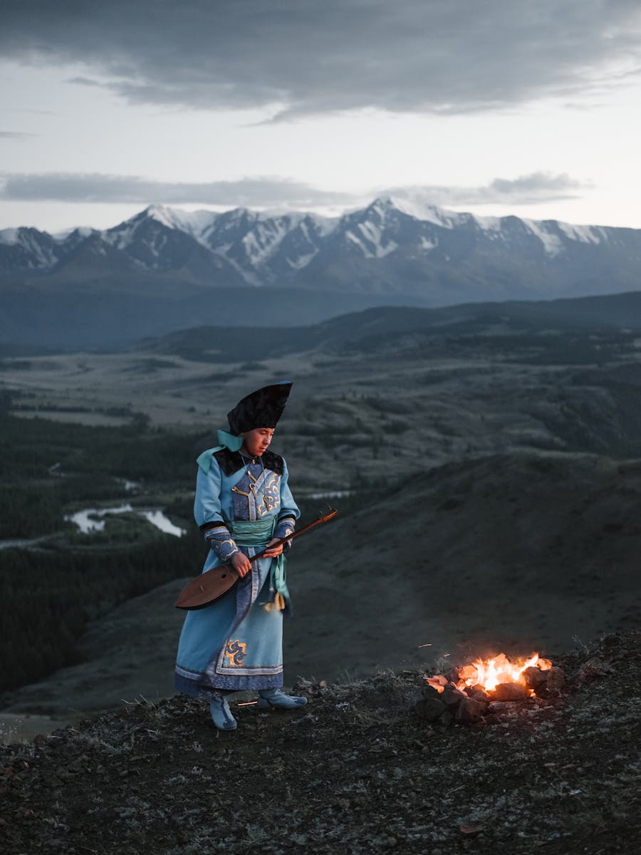 Full length of serious Tibetan male in traditional wear looking at campfire while standing with lute against mountains with cloudy sky at sunset